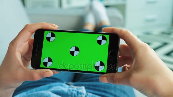 Mujer joven usando un teléfono inteligente negro con pantalla verde. Mujer sosteniendo el teléfono móvil en posición horizontal mientras está acostada en el sofá de la sala de estar. Clave de croma. Vista superior. Movimiento de rastreo. de cerca — Vídeo de stock