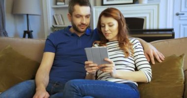 Mutlu genç çift kanepede oturan ve touchpad tablet cihaz üzerinde seyir. Tablet bilgisayar ile oturma odasında kanepede oturan aile