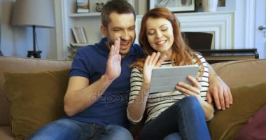Mutlu genç aile oturan kanepe oturma odasında, tablet cihaza kayıt üzerinde. Video sohbet arkadaşlarınızla konuşurken erkek ve kadın online video sohbet, skyping. Ellerini sallayarak
