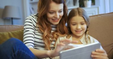 Tablet bilgisayar kullanarak kızı çekici annesiyle. Anne ve oturma odasında kanepede oturan ve tablet cihaz ile oynayan kız.