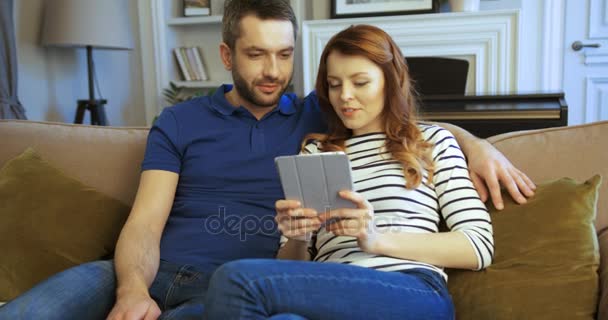 Счастливая молодая пара сидит на диване и смотрит на тачпад планшетного устройства. Семья сидит на диване в гостиной с планшетным компьютером — стоковое видео