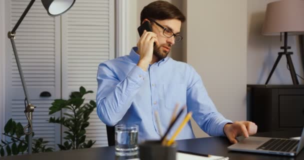 Un tânăr atrăgător vorbeşte la telefon în birou. Un tânăr care lucrează la laptop în birou . — Videoclip de stoc