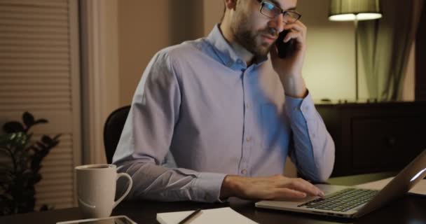 Attraktiver junger Mann, der im Büro mit dem Handy telefoniert. junger Mann, der im Büro am Laptop arbeitet. Spätdienst — Stockvideo