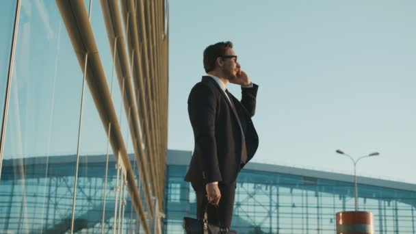 Atractivo hombre de negocios feliz hablando por teléfono mientras está de pie cerca del edificio de oficinas con grandes ventanas. Hombre sosteniendo el maletín. Lado wiew . — Vídeos de Stock