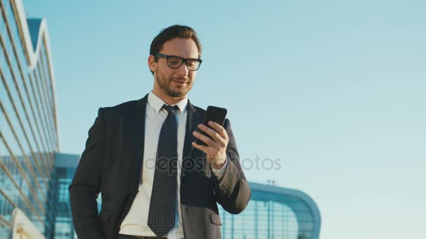 Atractivo hombre de negocios caucásico usando un teléfono inteligente mientras está parado fuera del edificio del centro de oficinas. Hombre leyendo, escribiendo en el teléfono celular y mirando a la cámara feliz. De cerca. — Vídeo de stock