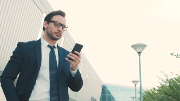 Jovem empresário atraente usando smartphone enquanto está ao ar livre perto do prédio do centro de escritórios. Homem a ler, a enviar mensagens no telemóvel. Fechar — Vídeo de Stock