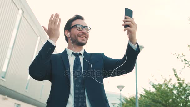 Atraente homem de negócios vídeo cahtting no smartphone fora perto do prédio do centro de escritórios. Homem a fazer videochamadas com colegas, família. Fechar — Vídeo de Stock