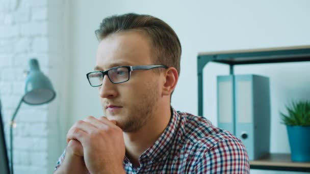 Retrato del atractivo hombre de negocios en las gafas pensando en algo que no sea volverse hacia la cámara, sonriendo y posando en la cámara mientras se sienta a la mesa en la oficina . — Vídeos de Stock