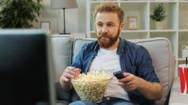 Genç adam anahtarlama kanalları TV Filmler ilginç bulmak, sit daha rahat ve başlangıç yemek ve patlamış mısır oturma odasında.
