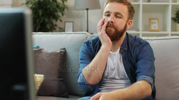 Atractivo hombre con barba se siente aburrido y tratando de encontrar una película interesante mientras está sentado en el sofá en la sala de estar . — Vídeos de Stock