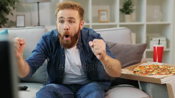 Молодой человек в повседневной рубашке ест пиццу и смотрит очень интересный футбольный матч по телевизору, сидя дома в гостиной . — стоковое видео
