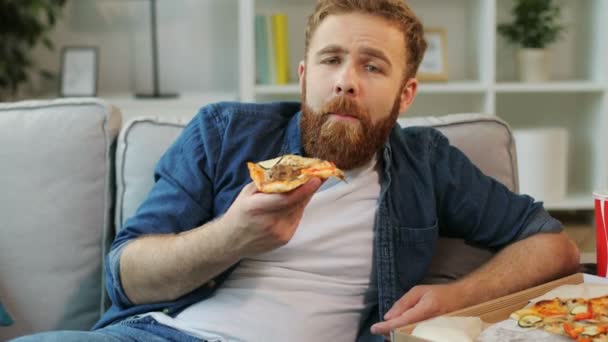 Portrét mladého bokovky s vousy, jíst pizzu při sledování sériových v televizi, zatímco sedí doma v obývacím pokoji. Detailní záběr. — Stock video