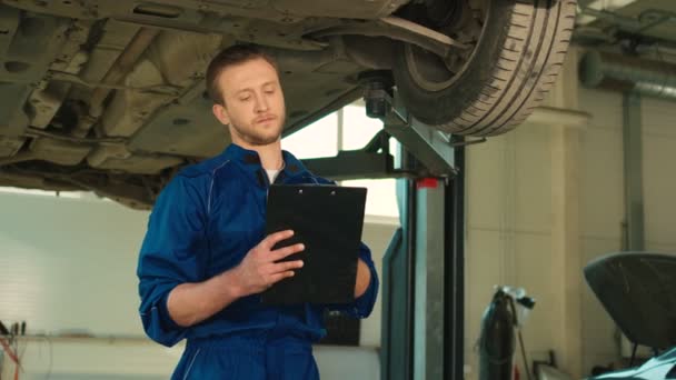 Koncentrerad kaukasiska auto mekaniker mannen autoservice att göra beskrivningen till anteckningsboken om tillståndet i bil hjulen. Inomhus. — Stockvideo