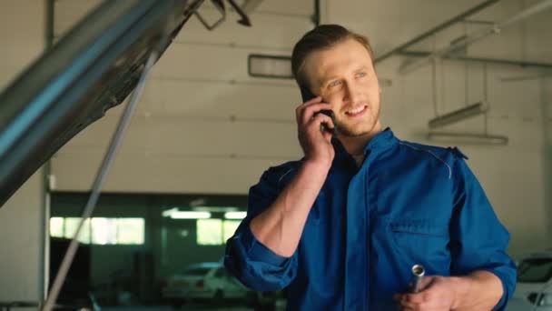Kaukasischer junger Automechaniker in blauer Uniform, der auf dem Smartphone mit einem Freund spricht, der im Autoservice steht und einen Steckschlüssel in der Hand hält. Innenräume. — Stockvideo