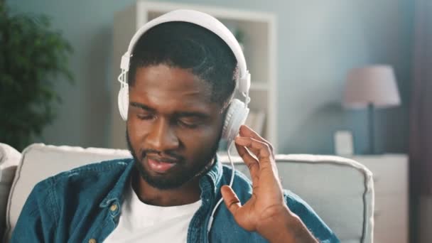 Schwarzer afroamerikanischer Mann mit weißen Kopfhörern, der zu Hause Musik auf seinem Smartphone hört. Zeitlupe — Stockvideo