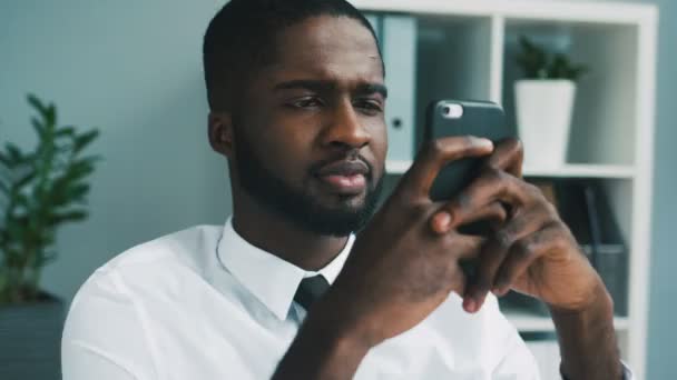 Retrato del hombre de negocios africano enviando mensajes en la oficina. Hombre africano joven sentado en la oficina y usando el teléfono inteligente . — Vídeo de stock