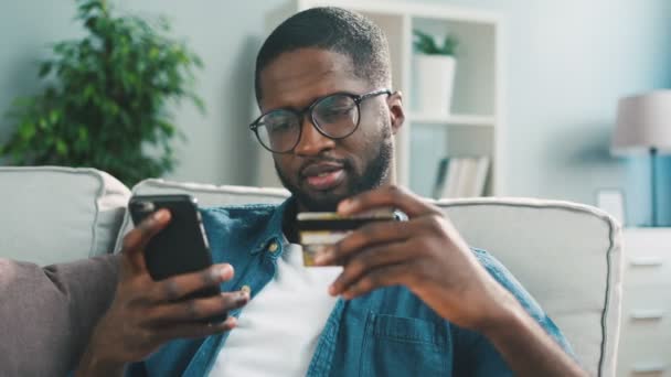 African young man in glasses shopping online with credit card using smart phone at home. Indoor. — Stock Video