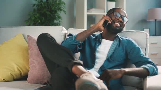 Vonzó afrikai férfi mennyezeten a telefon, a nappaliban a kanapén ülve. Fiatal ember használ szúró telefon chating. — Stock videók