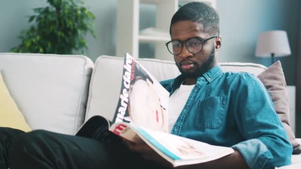 Jovem africano com barba em camisa azul sentado no sofá branco e revista de visualização concentrada para fazer compras. Interior . — Vídeo de Stock