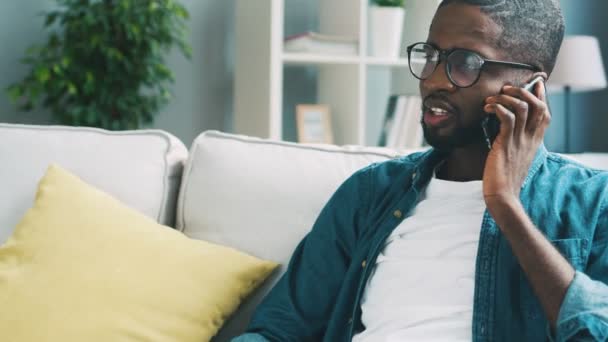 Joven hombre africano en gafas sentado en el sofá hablando a través de un teléfono inteligente mientras usa la tableta. Hombre africano sonriendo cuando se utiliza el teléfono inteligente . — Vídeo de stock