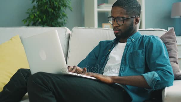 Attraktiv afrikanska man i glas som sitter i soffan med laptop hemma på levande bakgrund för chatta med vänner. Inomhus. — Stockvideo