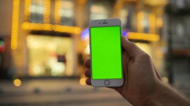 Akşam şehir sokak arka plan üzerinde beyaz smartphone ile yeşil ekran holding erkek eller yukarı kapatın. Güzel ışıkları ile bina. Yakın çekim. Chroma anahtar