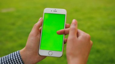 Yeşil çim zemin üzerine beyaz smartphone ile yeşil ekran tutan el. Park açık havada. Kaydırma, yakınlaştırma, dokunmatik ekran dokunarak kadın. Chroma anahtar. Yakın çekim.
