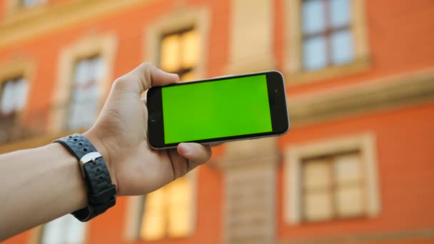남자 손을 잡고 녹색 스크린 스마트폰 야외 도시에서 거리를 닫습니다. 수평. 주황색 건물 배경입니다. 크로마 키 — 비디오