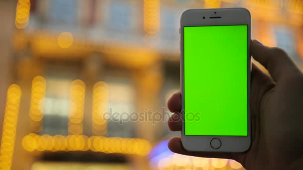 Mani maschili in possesso di smartphone bianco con schermo verde sullo sfondo della strada della città nella notte. Bellissimo edificio con luci. Chiudete. Chiave cromatica — Video Stock