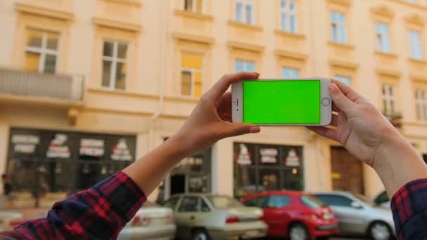 Женщины держат в руках белый смартфон с зеленым экраном на открытом воздухе на фоне городской улицы. Горизонтально. Хрома-ключ. Закрыть — стоковое видео