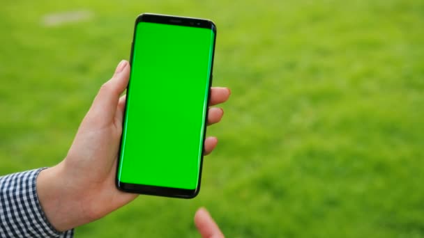 Mão feminina segurando smartphone preto com tela verde no fundo de grama verde. Ao ar livre no parque. Mulher rolando, tocando no touchscreen. Chave Chroma. Fecha. Movimento de rastreamento — Vídeo de Stock