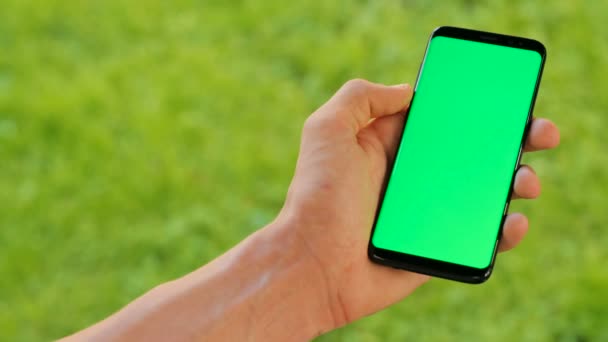 Mužské ruky držící černý smartphone s zeleným plátnem na zelené trávě pozadí. Venku v parku. Chromatický klíč. Detailní záběr. — Stock video