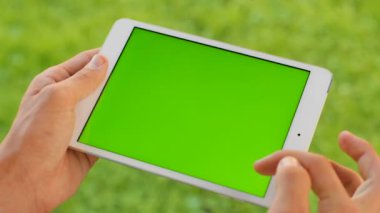 Yeşil çim zemin üzerine beyaz tablet bilgisayar cihaz ile yeşil perde tutan adam. Park açık havada. Kaydırma, yakınlaştırma, dokunmatik ekran dokunarak bir adam. Chroma anahtar. Yakın çekim.