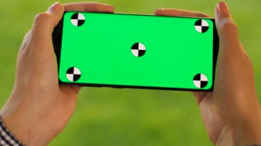 Yeşil çim zemin üzerine siyah smartphone ile yeşil ekran tutan el. Park açık havada. Chroma anahtar. Yakın çekim. Yatay. Hareket izleme