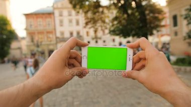 Güneşli günde şehir sokak arka plan üzerinde beyaz akıllı telefon ile yeşil perde tutan adam. Yatay. Chroma anahtar. Yakın çekim