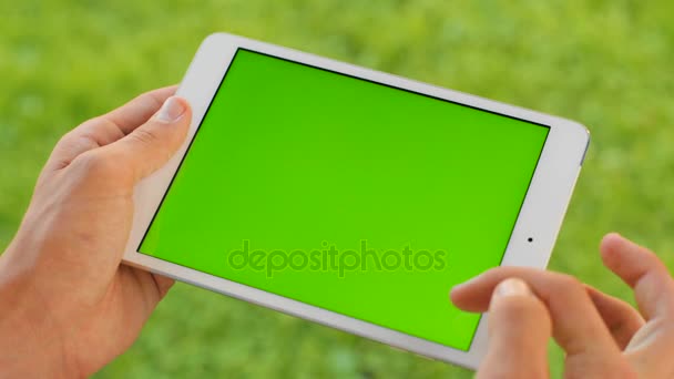 Чоловік тримає білий планшетний комп'ютерний пристрій з зеленим екраном на фоні зеленої трави. На відкритому повітрі в парку. Чоловік прокручує, збільшує, натискає на сенсорний екран. Ключ хроми. крупним планом . — стокове відео