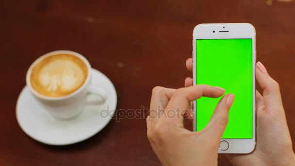 Female hands holding white smartphone with green screen on wooden backgroung in the cafe. Woman scrolling, tapping, zooming and drinking coffee. Close up. Chroma key — Stock Video