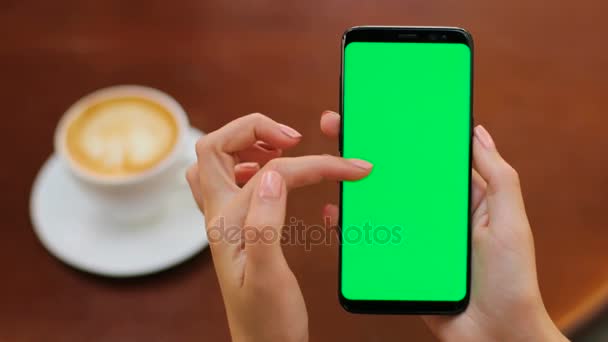 Mãos femininas segurando smartphone preto com tela verde no backgroung de madeira no café. Mulher rolando, tocando, ampliando. Fecha. Chave Chroma — Vídeo de Stock