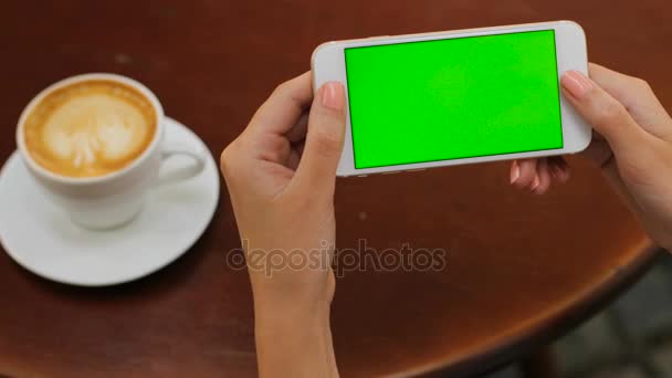 Ženské ruce držící bílý smartphone s zeleným plátnem na dřevěné backgroung v kavárně. Vodorovně. Žena, pití kávy. Zblízka. Chroma klíč