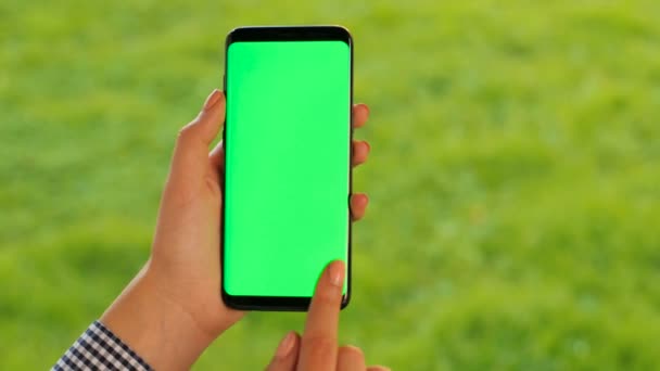 Mano femenina sosteniendo teléfono inteligente negro con pantalla verde sobre fondo de hierba verde. Al aire libre en el parque. Mujer desplazándose, tocando en la pantalla táctil. Clave de croma. De cerca. . — Vídeos de Stock