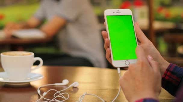 Manos femeninas sosteniendo teléfono inteligente blanco con pantalla verde y auriculares mientras se sienta al aire libre en la cafetería. Mujer desplazándose. zoom en la pantalla táctil. Vertical. De cerca. Clave de croma — Vídeo de stock