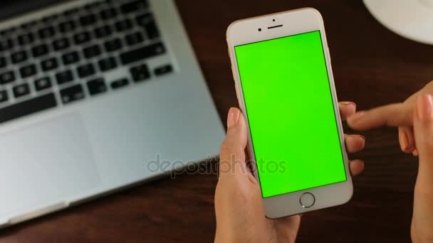 Female hands holding smart phone with green screen on wooden table background with laptop keyboard. Woman scrolling, zooming. Close up. Chroma key — Stock Video