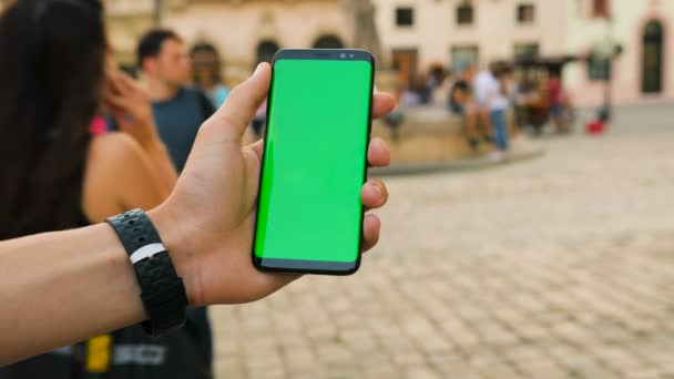 Homem segurando telefone inteligente preto com tela verde no fundo da rua da cidade no dia ensolarado. Um grande ecrã. Chave Chroma. Fechar — Vídeo de Stock