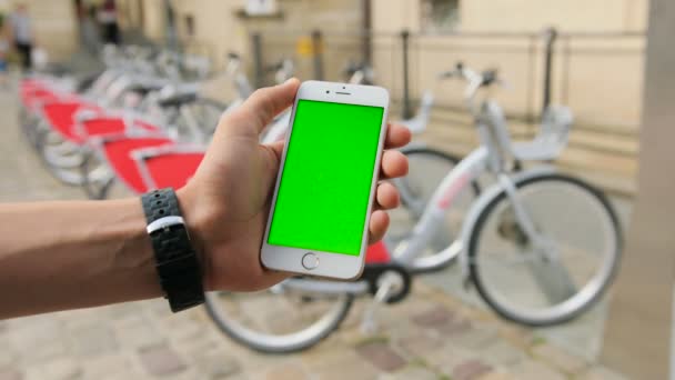 Αρσενικό χέρια κρατώντας λευκό smartphone με πράσινη οθόνη street φόντο πόλη. Ποδήλατο επιτόπου. Υπηρεσία ενοικίασης ποδηλάτων. Νοικιάστε ένα ποδήλατο. Κλειδί Chroma. Κοντινό πλάνο — Αρχείο Βίντεο