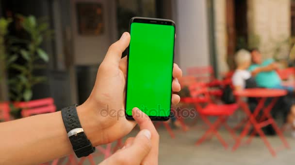 Male hands holding black smartphone with green screen on street cafe background. Man scrolling, swiping pictures. Close up. Chroma key. — Stock Video