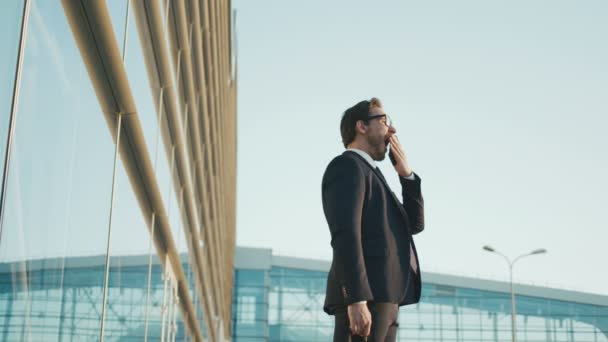 Молодой стильный бизнесмен стоит снаружи офисного здания, зевая и растягивая . — стоковое видео