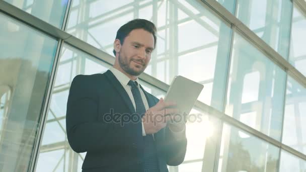 Attractive business man using tablet device, surfing internet, reading, textin on digital tablet device. Outdoor shot near office center building. Close up — Stock Video