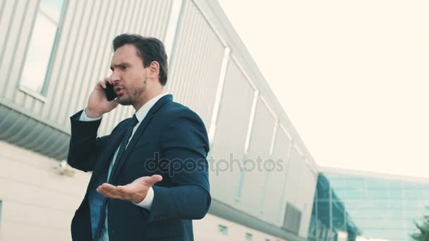 Молодой бизнесмен чувствует себя раздраженным, разговаривая по мобильному телефону на улице возле офисного здания центра. Человек злой и грустный. Закрыть — стоковое видео