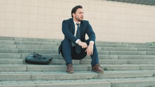 Der junge Geschäftsmann sitzt auf der Treppe in der Nähe von Bürogebäuden und fühlt sich schlecht, traurig und genervt. Mann müde und mit Problemen, die Hände auf den Kopf legen — Stockvideo