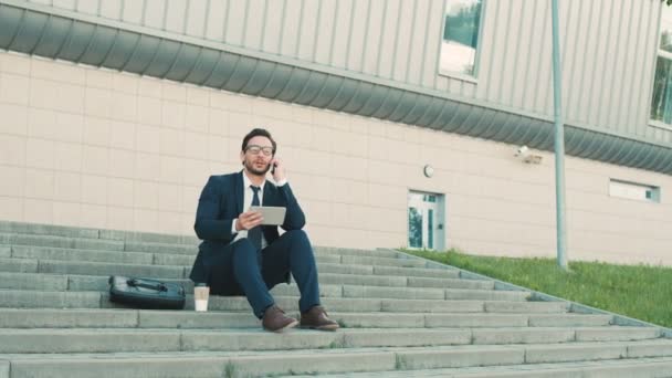 Ung snygg affärsman sitter på trappan nära kontorsbyggnad, använda digitala surfplatta dela, talar på mobilen och dricka kaffe. — Stockvideo
